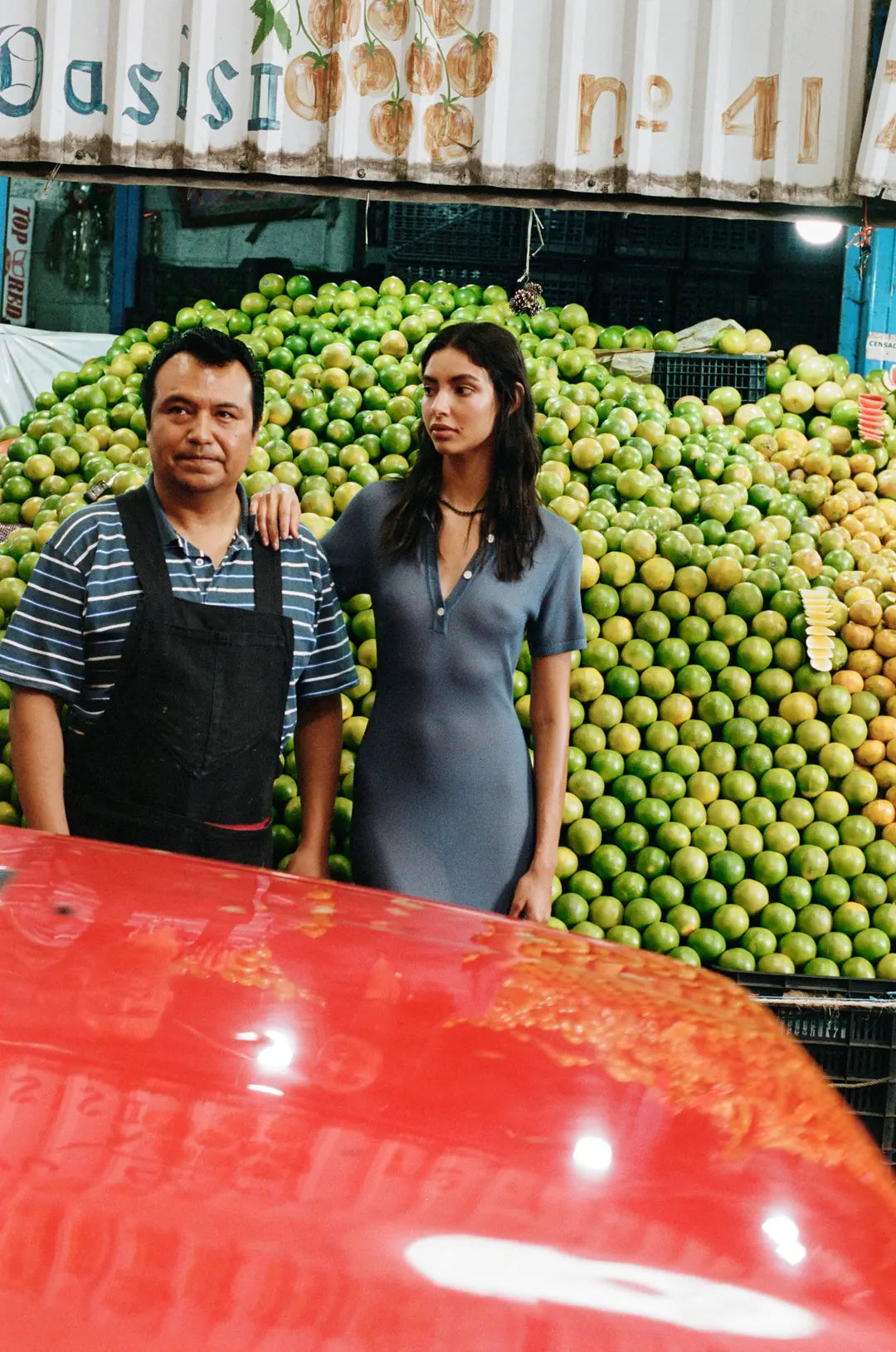 Two people standing in front of a large pile of green apples with a red vehicle