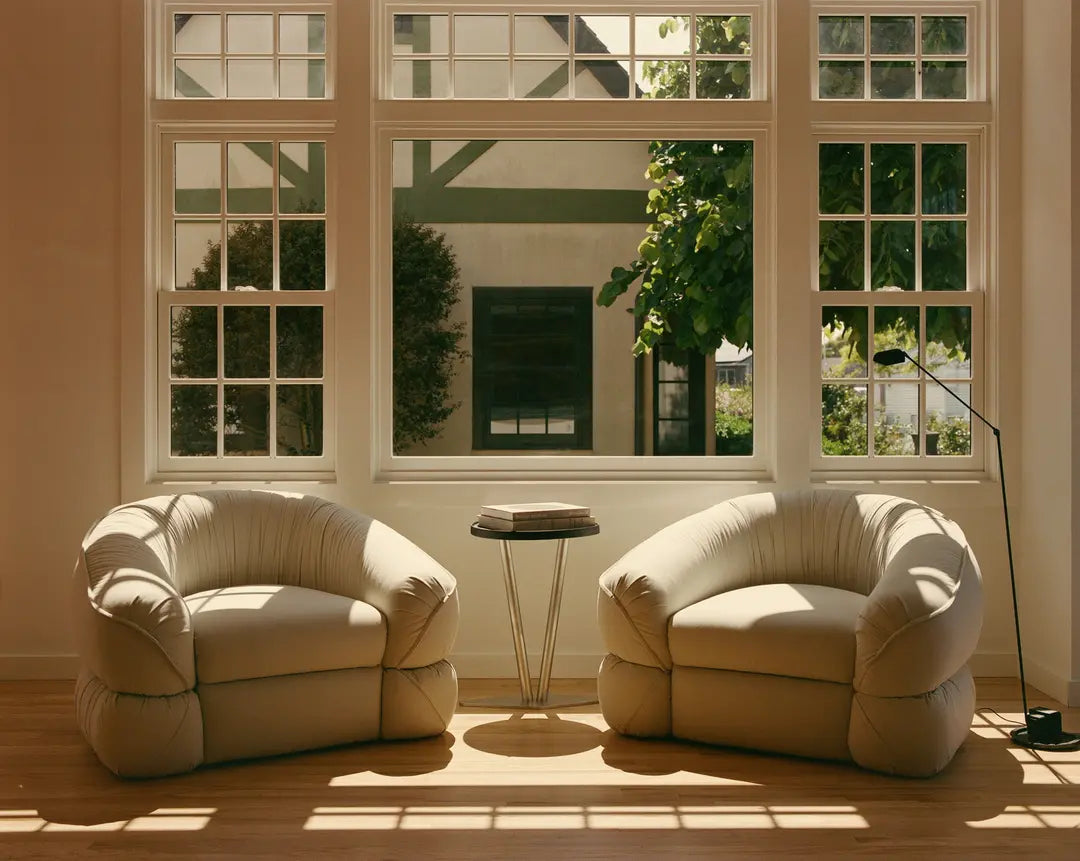 Two beige armchairs in a room with large windows and a small table